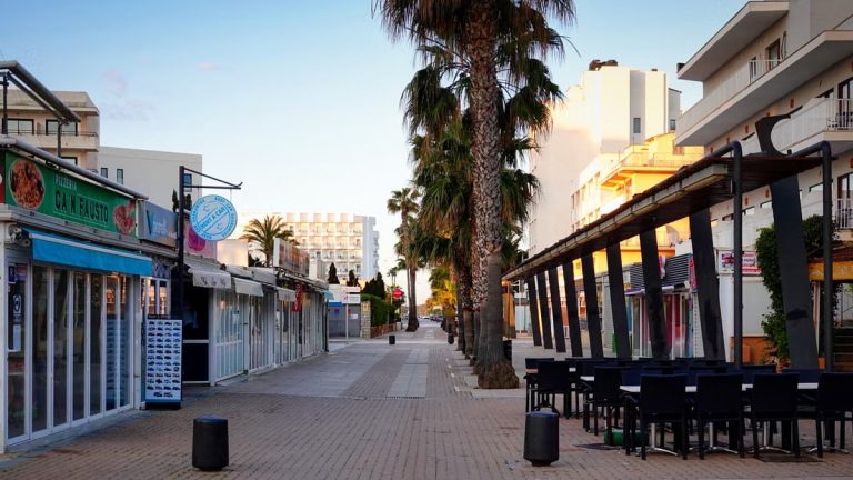 A peaceful, sunlit street in Cala Millor, Spain with palm trees and buildings lining the path.