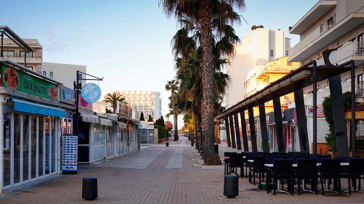 A peaceful, sunlit street in Cala Millor, Spain with palm trees and buildings lining the path.